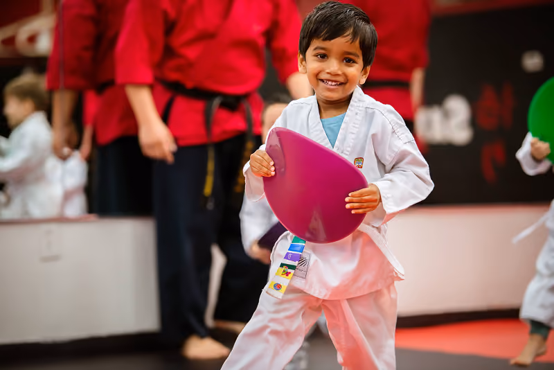 Smiling young boy in a white martial arts uniform holding a purple training pad in a dojo with other children and instructors wearing red uniforms in the background.