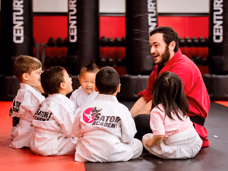 A martial arts instructor in a red gi talks with five young children wearing white martial arts uniforms, sitting on a gym floor with punching bags in the background.