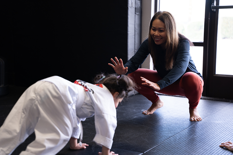 Woman in a black long sleeve shirt and red leggings squats smiling with hands outstretched toward a child in a white martial arts uniform who is on hands and feet on a mat.