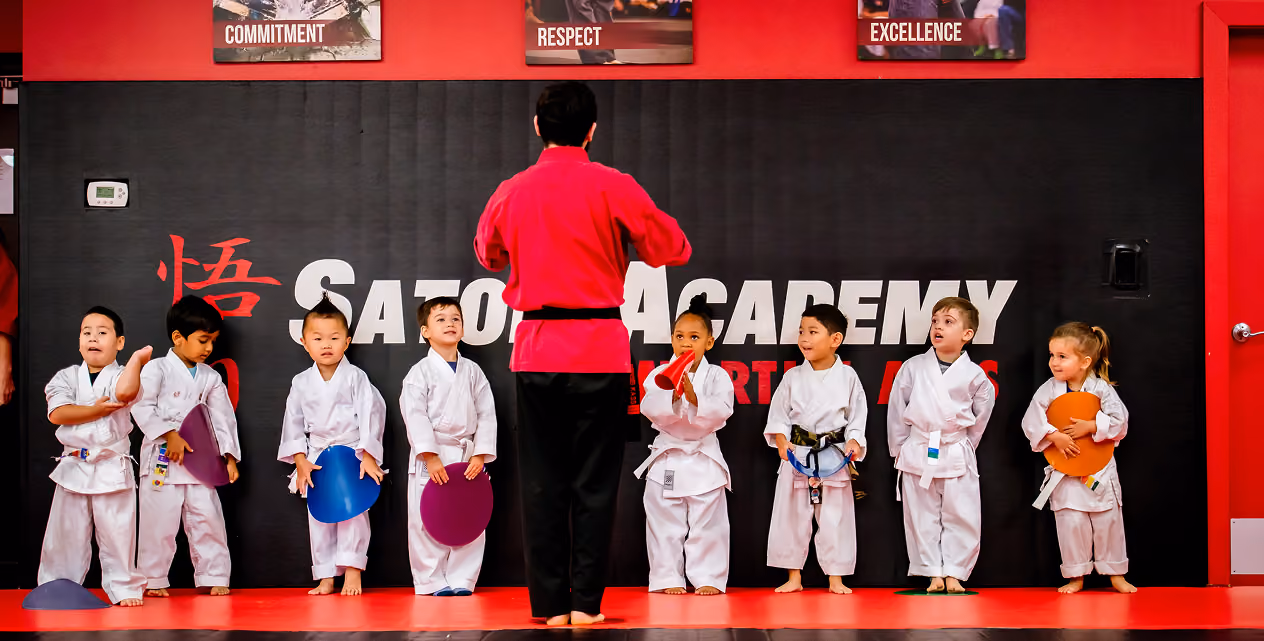Martial arts instructor in red uniform facing a line of eight children in white gis holding colored kicking pads inside Satori Academy with red and black walls and motivational banners.