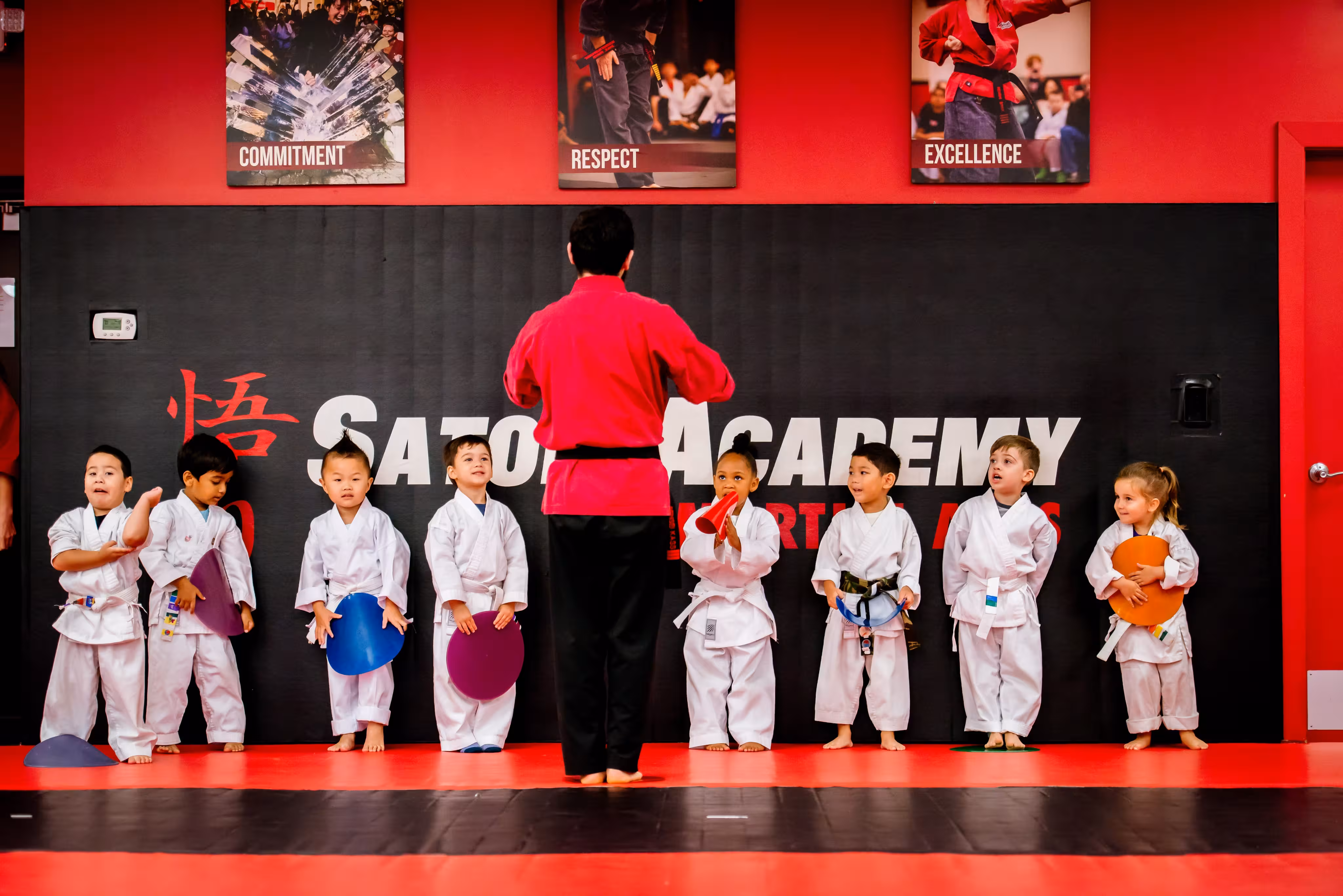 Martial arts instructor in a red uniform stands barefoot facing a line of eight young children in white karate uniforms holding colored kicking targets inside a dojo with red and black mats and a black wall with the text 'Satori Academy' and posters reading Commitment, Respect, and Excellence above.