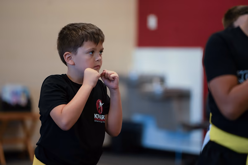 Young boy in a black martial arts uniform with a yellow belt, standing in a fighting stance with fists raised indoors.