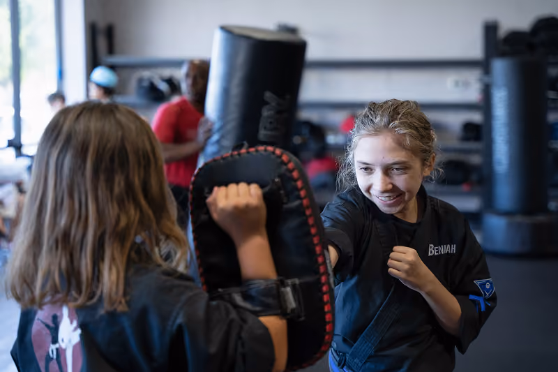 Two girls practicing martial arts in a dojo, one holding a padded target and the other, wearing a black uniform with the name Beniah, preparing to strike it.