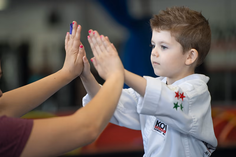Young boy in a martial arts uniform with star patches on the sleeve touches hands with an adult wearing a watch and with a purple bandage on one finger.
