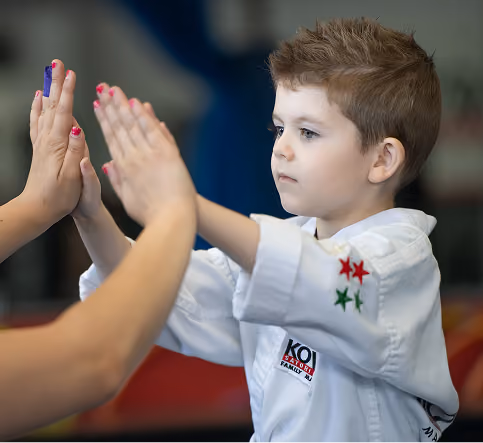 A young boy in a white martial arts uniform with star patches practices hand techniques facing an adult with painted nails in a training setting.