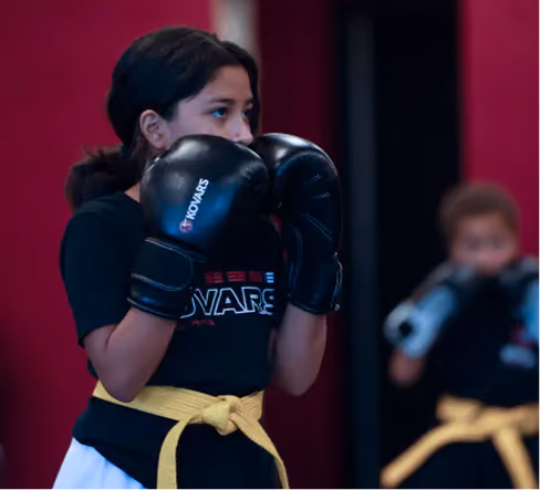 Young girl wearing black boxing gloves and a yellow belt practices boxing in a gym, with another child similarly equipped in the background.