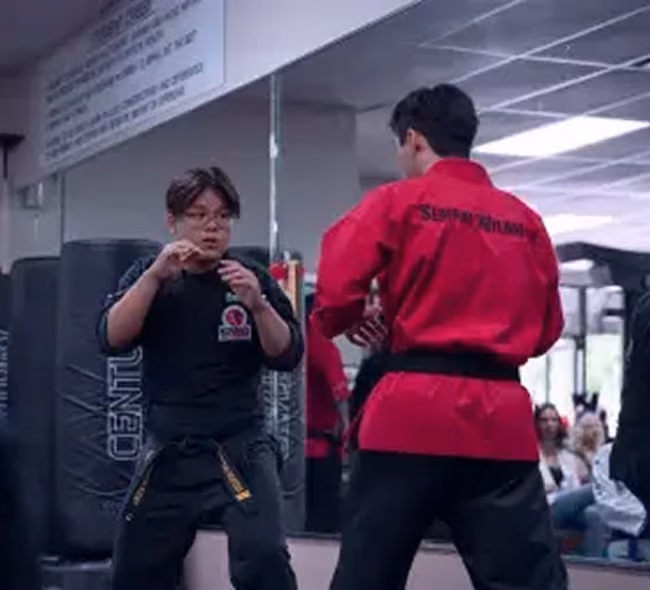 Two martial artists in a training room facing each other in fighting stance with one wearing a black uniform and the other wearing a red uniform with a black belt.