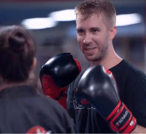 A man with short hair wearing black and red boxing gloves smiling and facing a woman in martial arts attire with her back to the camera, in an indoor training setting.