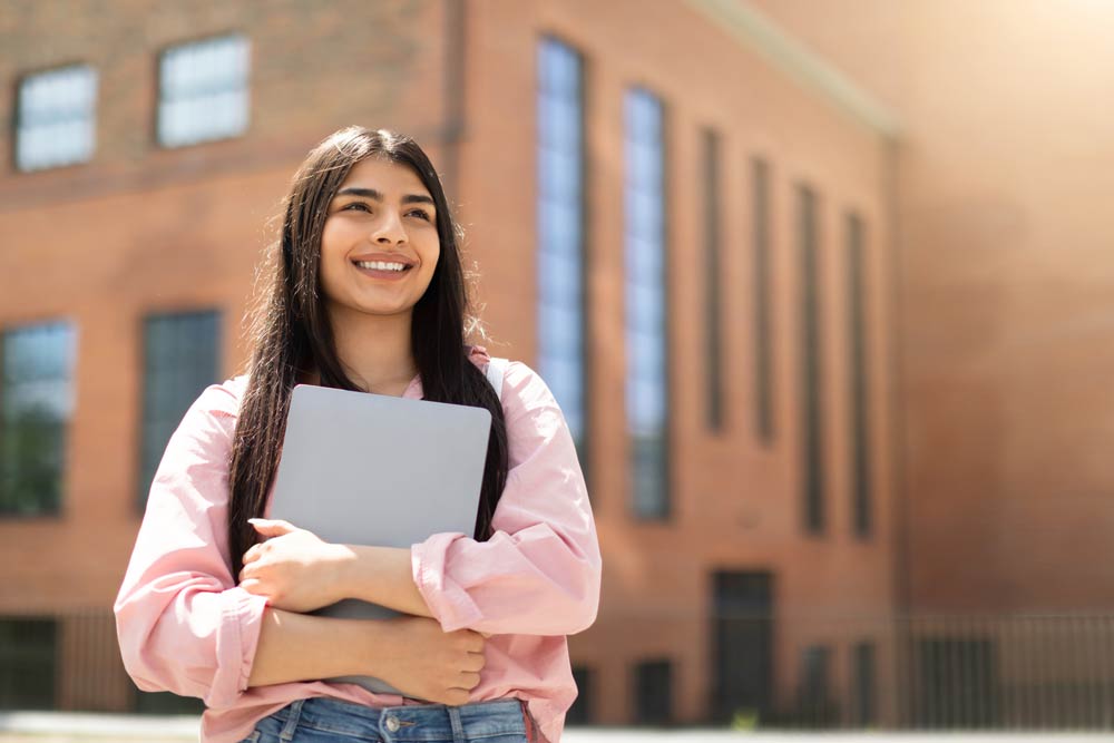 A smiling teenage student holding a laptop outside a school building, representing confidence and planning for post-high school options for teens with ADHD.