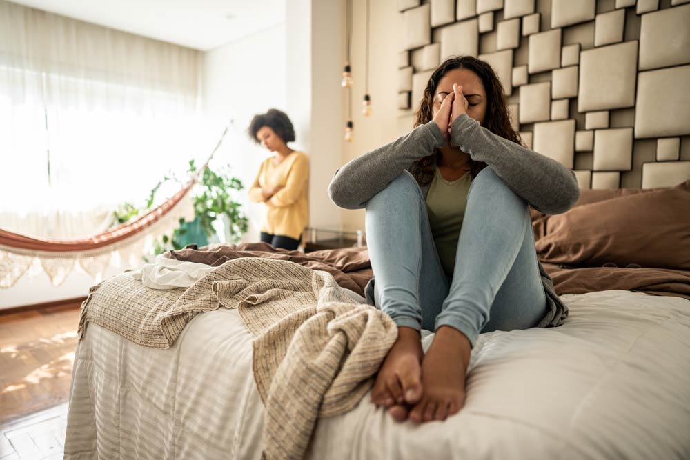 Couple in conflict, with one sitting on the bed holding her face in distress while the other stands with arms crossed, representing the challenges of managing impulsivity and emotional intensity in ADHD relationships.