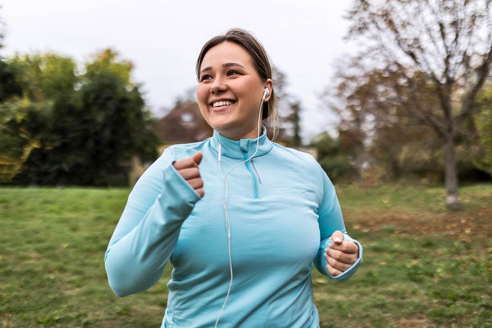 Smiling young woman jogging outdoors with headphones, representing how exercise supports focus, mood, and symptom management for individuals with ADHD.