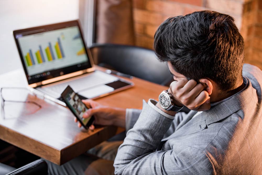 Man distracted by his phone while work charts display on a laptop, representing ADHD-related procrastination and task initiation challenges.