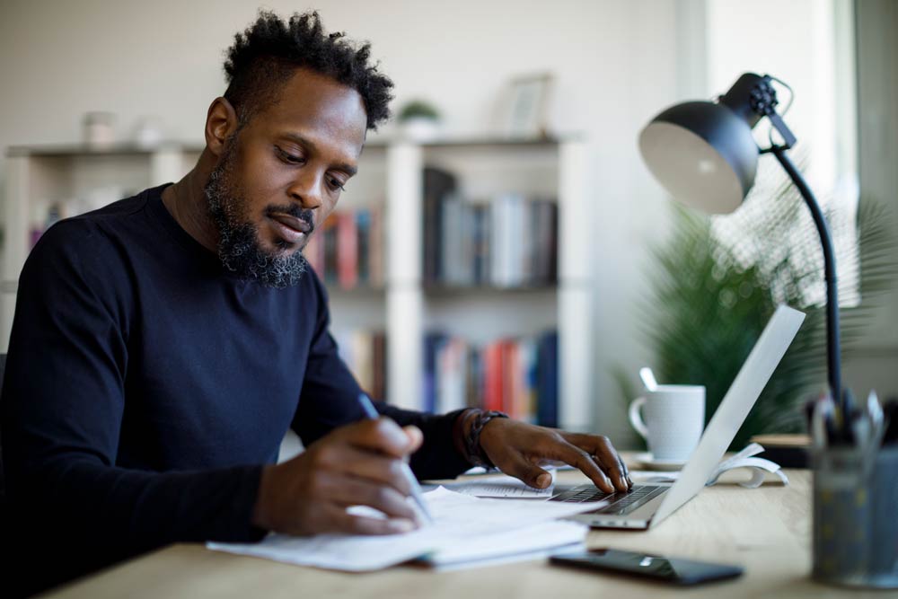 Man writing notes while working on a laptop at a desk, representing the use of ADHD assessment tools and diagnostic procedures.