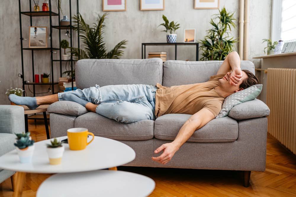 An adult lying tired on a couch with an arm over his eyes, representing inattentive ADHD symptoms, fatigue, and executive functioning challenges at home.