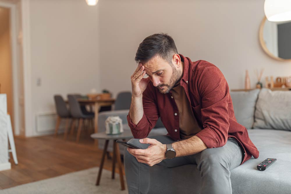 A man sitting on a couch looking concerned while reading his phone, representing frustration and emotional strain from delays in ADHD diagnosis and care.