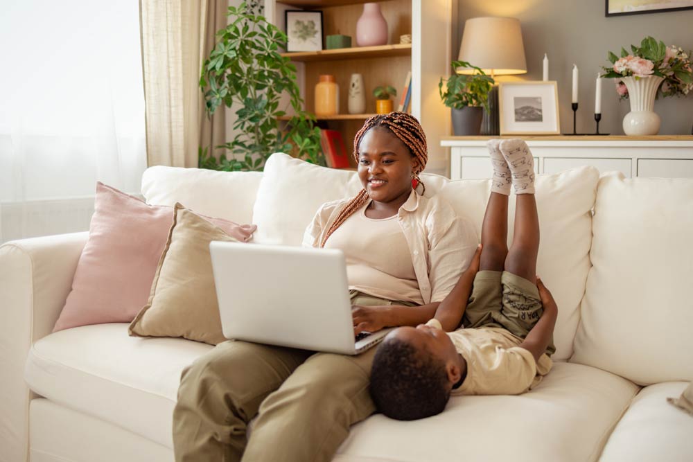 Mother with ADHD smiling while working on a laptop as her playful child lies beside her lap, representing joyful parenting and connection through humour.