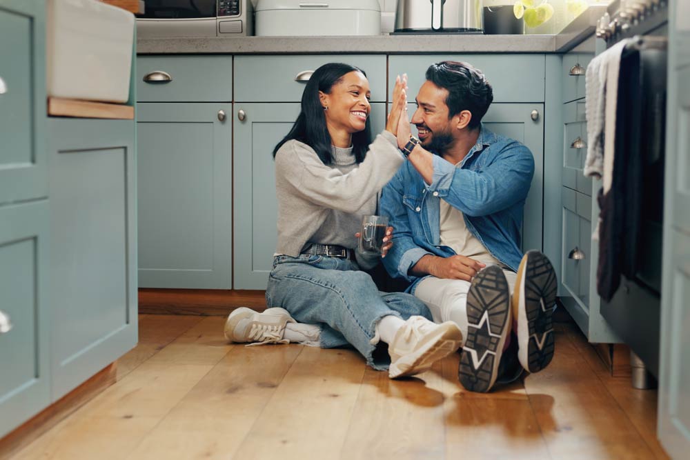 A smiling couple sitting on a kitchen floor giving each other a high five, representing teamwork, positive communication, and shared success in ADHD co-parenting.
