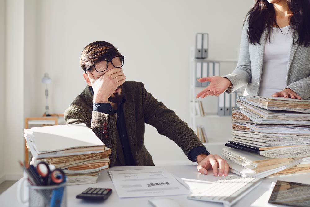 A man sitting at a cluttered desk with his hand over his face while a colleague gestures nearby, symbolizing ADHD-related forgetfulness, workplace stress, and executive function challenges.