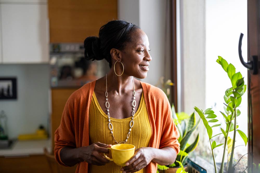 A woman smiling while holding a cup of tea and looking out a window, symbolizing calm reflection and balance in managing ADHD symptoms affected by hormonal changes.