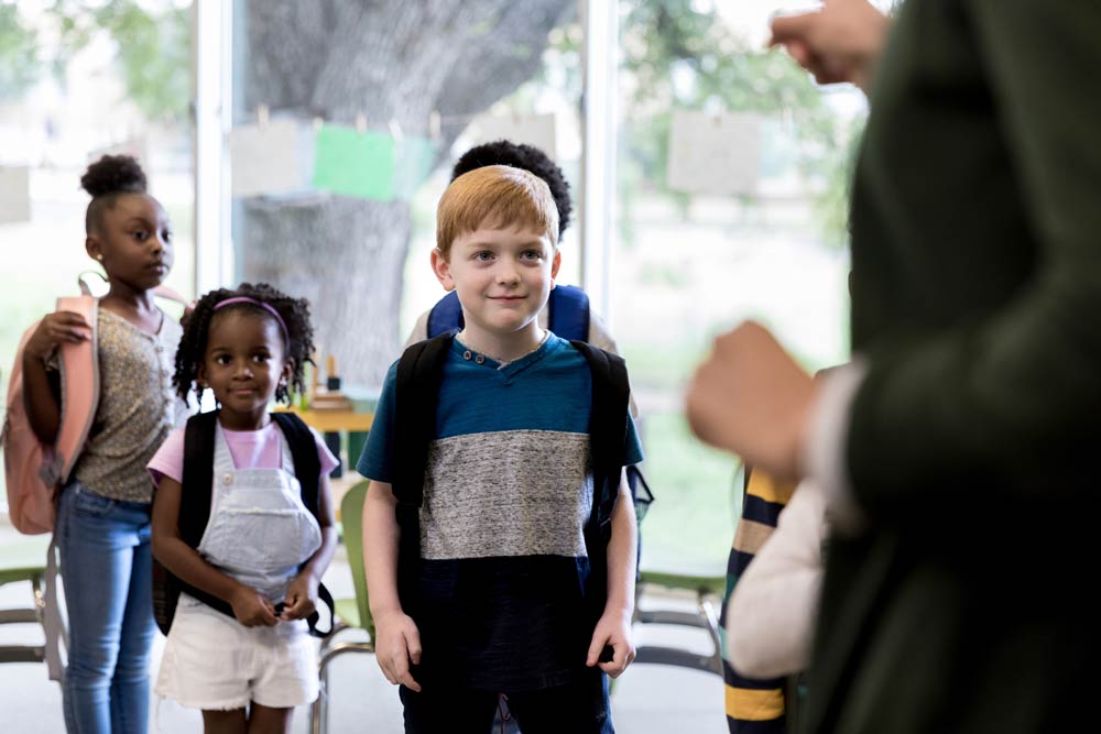 Children with backpacks line up in a classroom while listening to a teacher, illustrating how kids with ADHD can learn turn-taking and conversation skills through structured guidance.