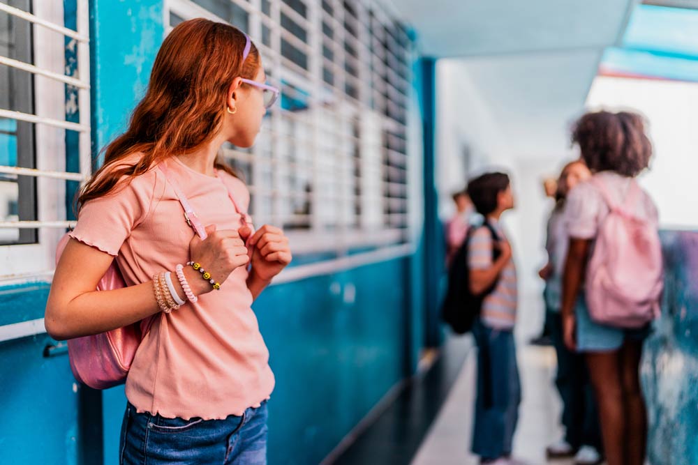 A young girl with a backpack looks on while other children talk together at school, illustrating the challenges of bullying and social rejection for kids with ADHD.