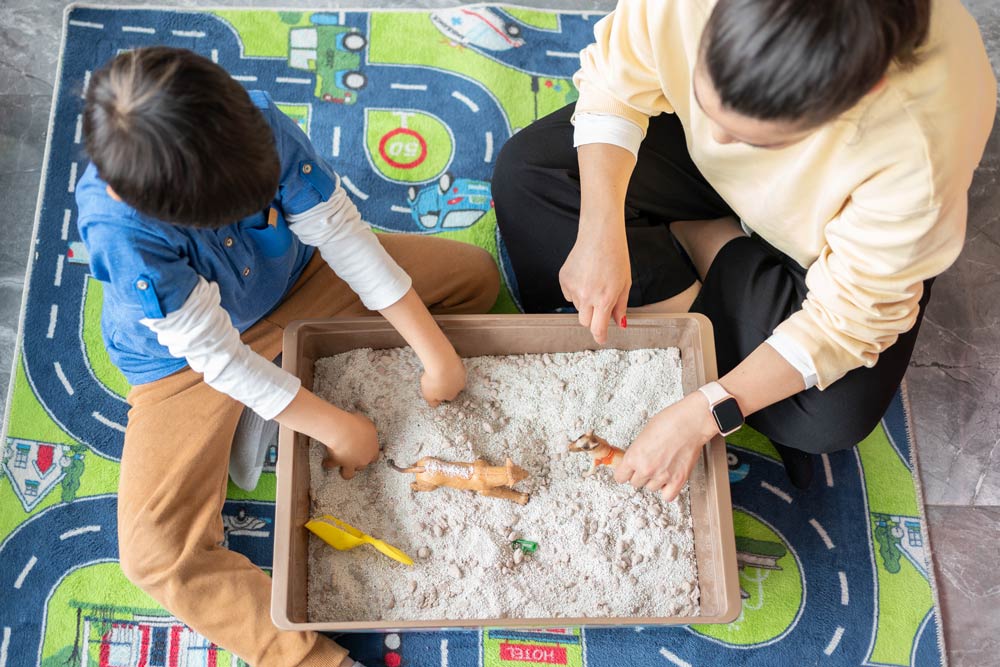 A young child and therapist play together in a sandbox during a session, illustrating how play therapy supports emotional regulation and social development for kids with ADHD.