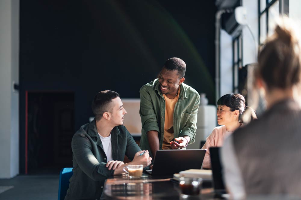 A group of people smiling and collaborating around a table with laptops, symbolizing the importance of social connection and teamwork in supporting ADHD well-being.