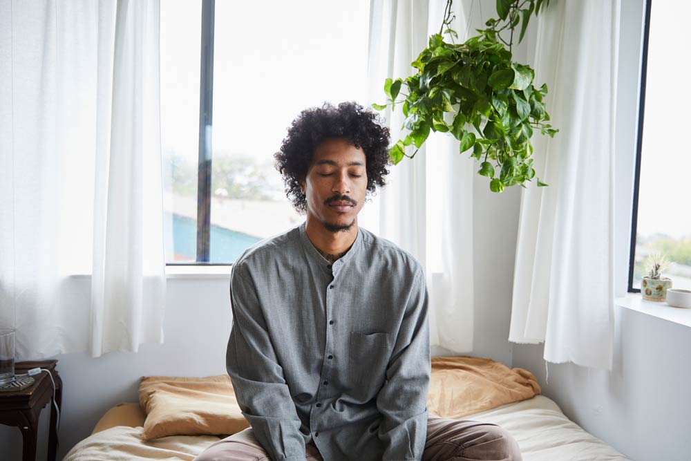 A man sitting cross-legged on a bed with eyes closed, practicing mindfulness and grounding to manage ADHD-related overwhelm and emotional stress.
