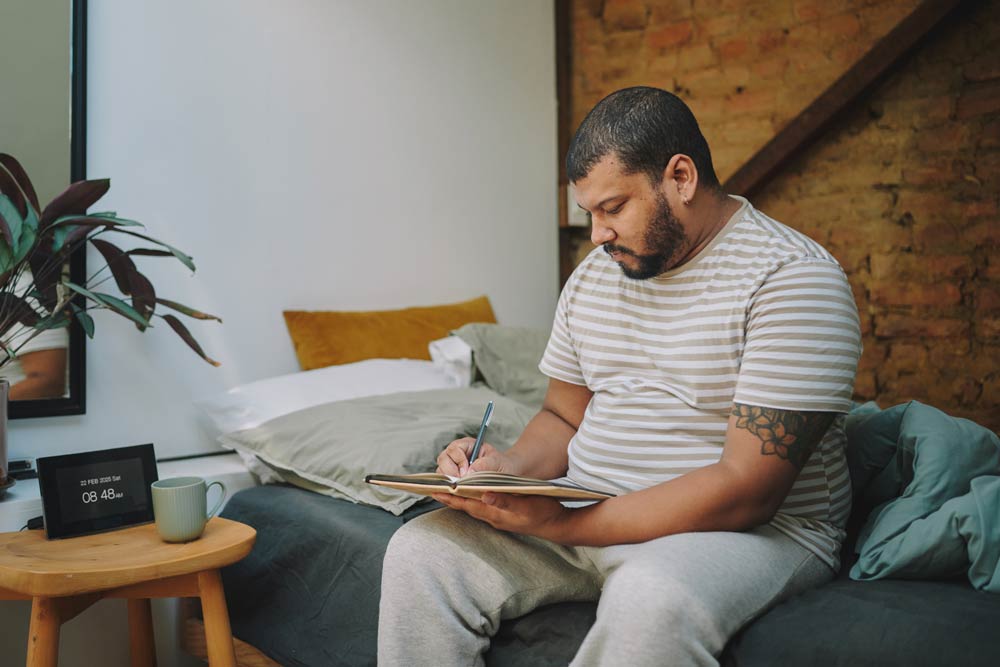 A man sitting on his bed writing in a notebook, representing how bullet journaling can help individuals with ADHD organize tasks, manage time, and improve focus.