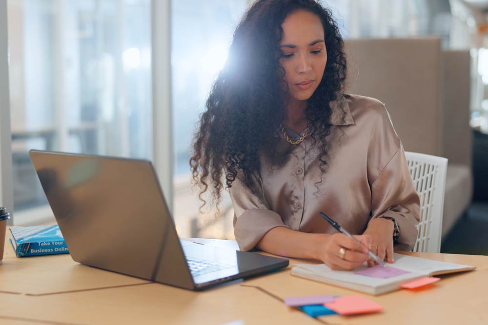 A woman sitting at her desk using both a laptop and a notebook, representing the comparison between digital and physical planners for managing ADHD tasks and improving organization.