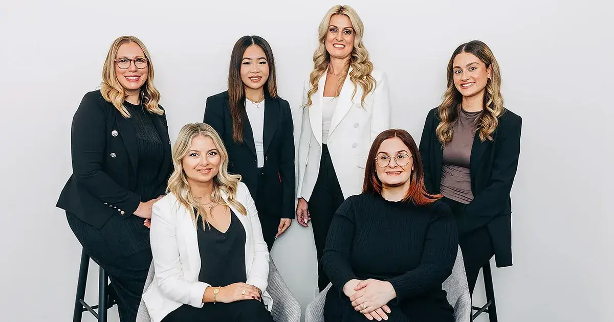 Group photo of six diverse women from the Moonee Valley Conveyancing team, five standing and one seated, dressed in professional business attire and smiling.
