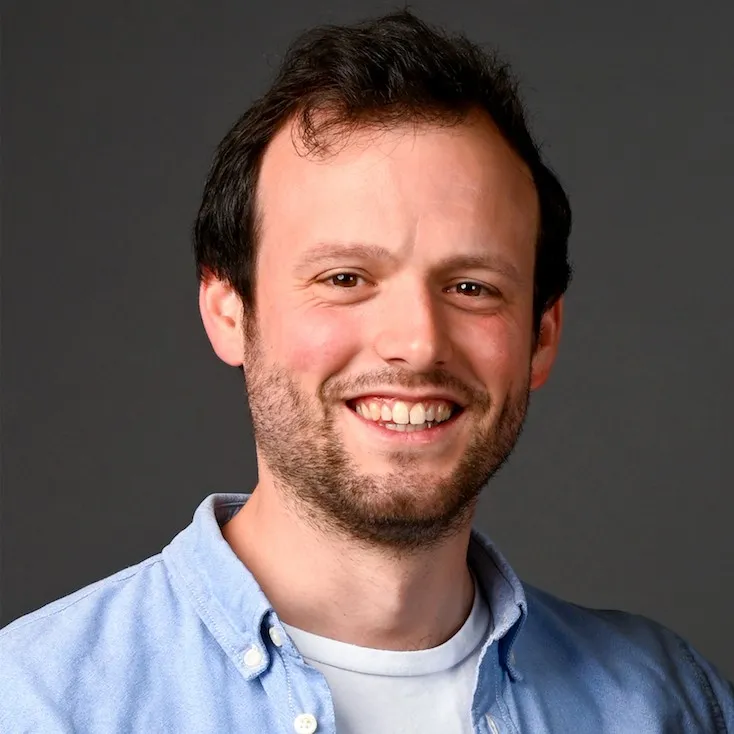 Smiling man with short dark hair, beard, and light blue button-up shirt over white t-shirt, against a dark gray background.