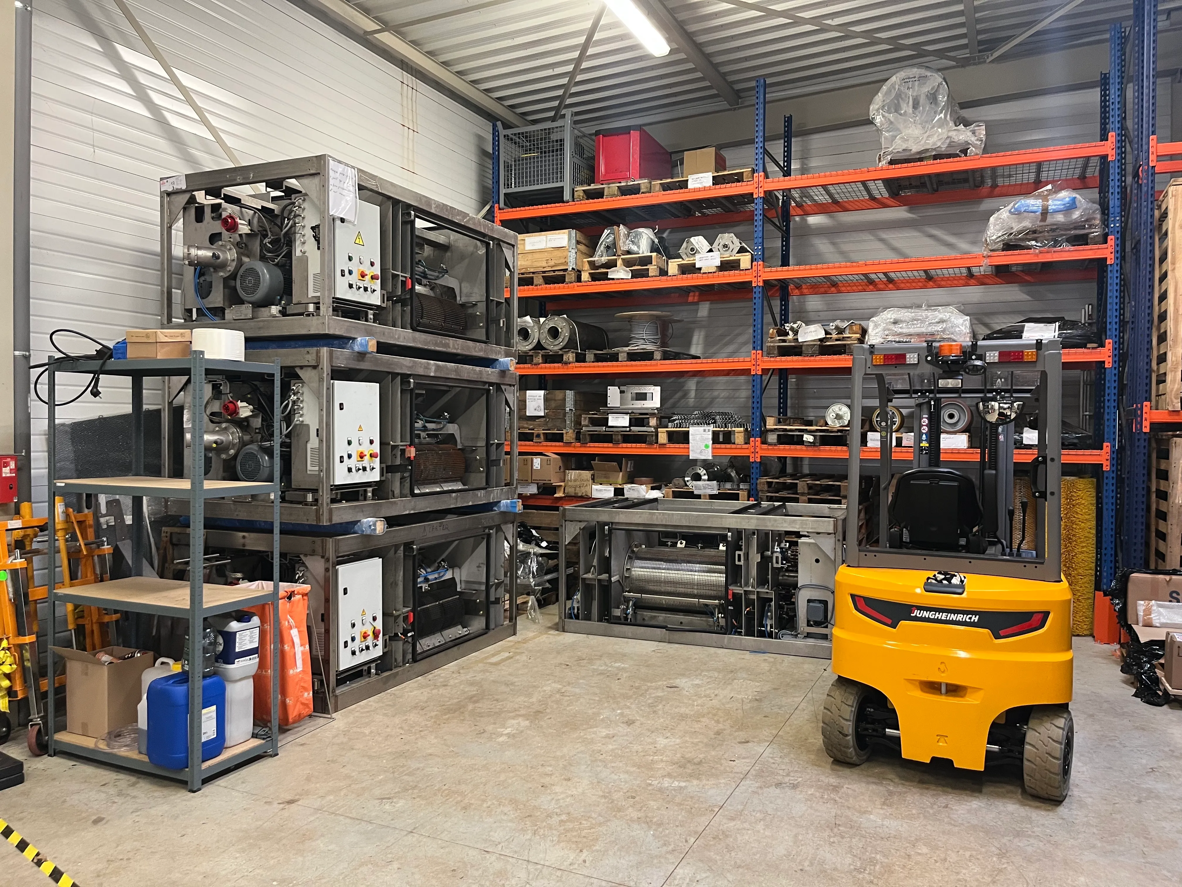Indoor warehouse with stacked industrial machinery, metal shelves holding boxes and parts, and a yellow Jungheinrich forklift in the foreground.