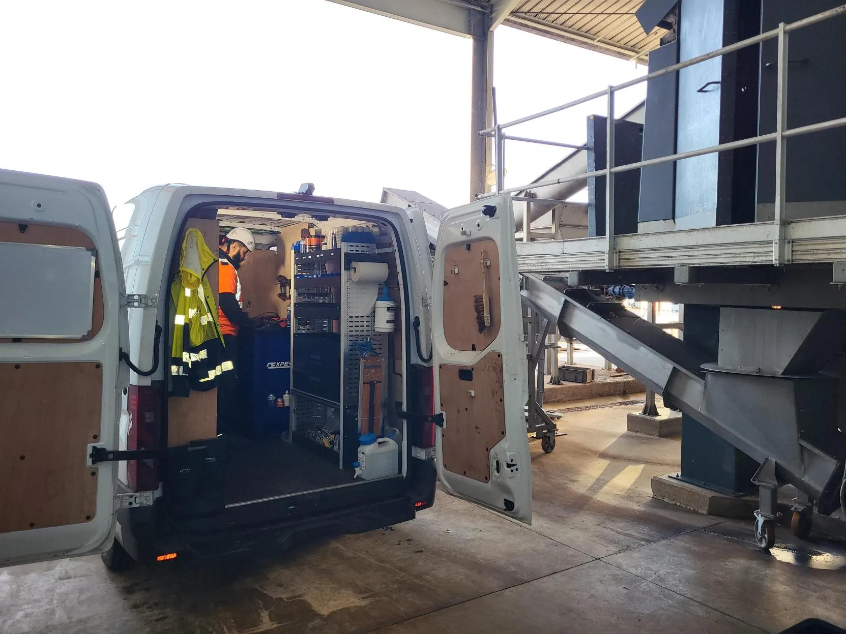 Worker in an orange safety vest and helmet standing inside a white van with open rear doors, equipped with organized shelves and tools, parked in an industrial setting next to metal machinery.