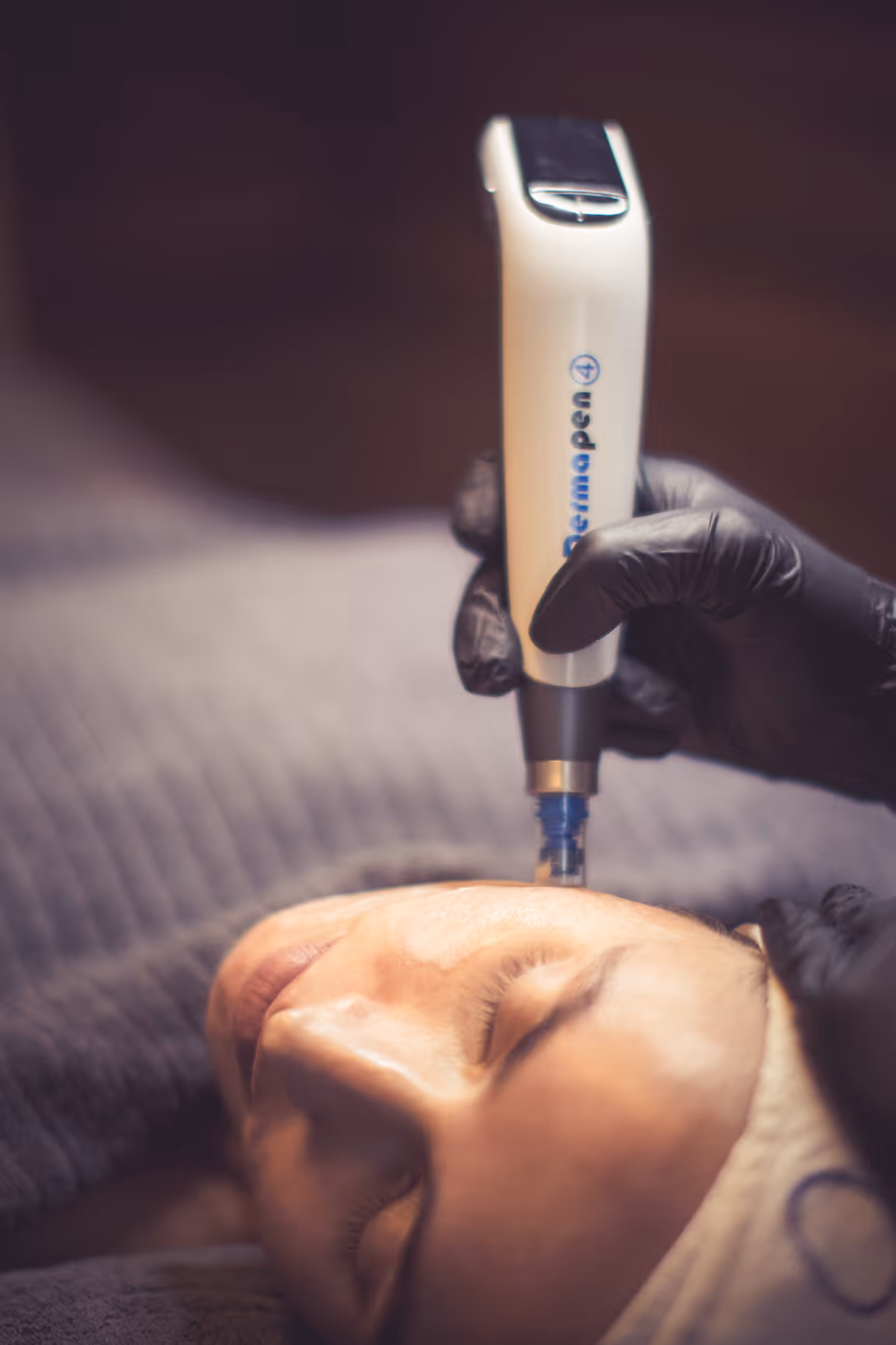 En person får en ansiktsbehandling med en HydraFacial-enhet som holdes i en hanskekledd hånd. Personen ligger avslappet på et behandlingsbord med lukkede øyne og et pannebånd på.