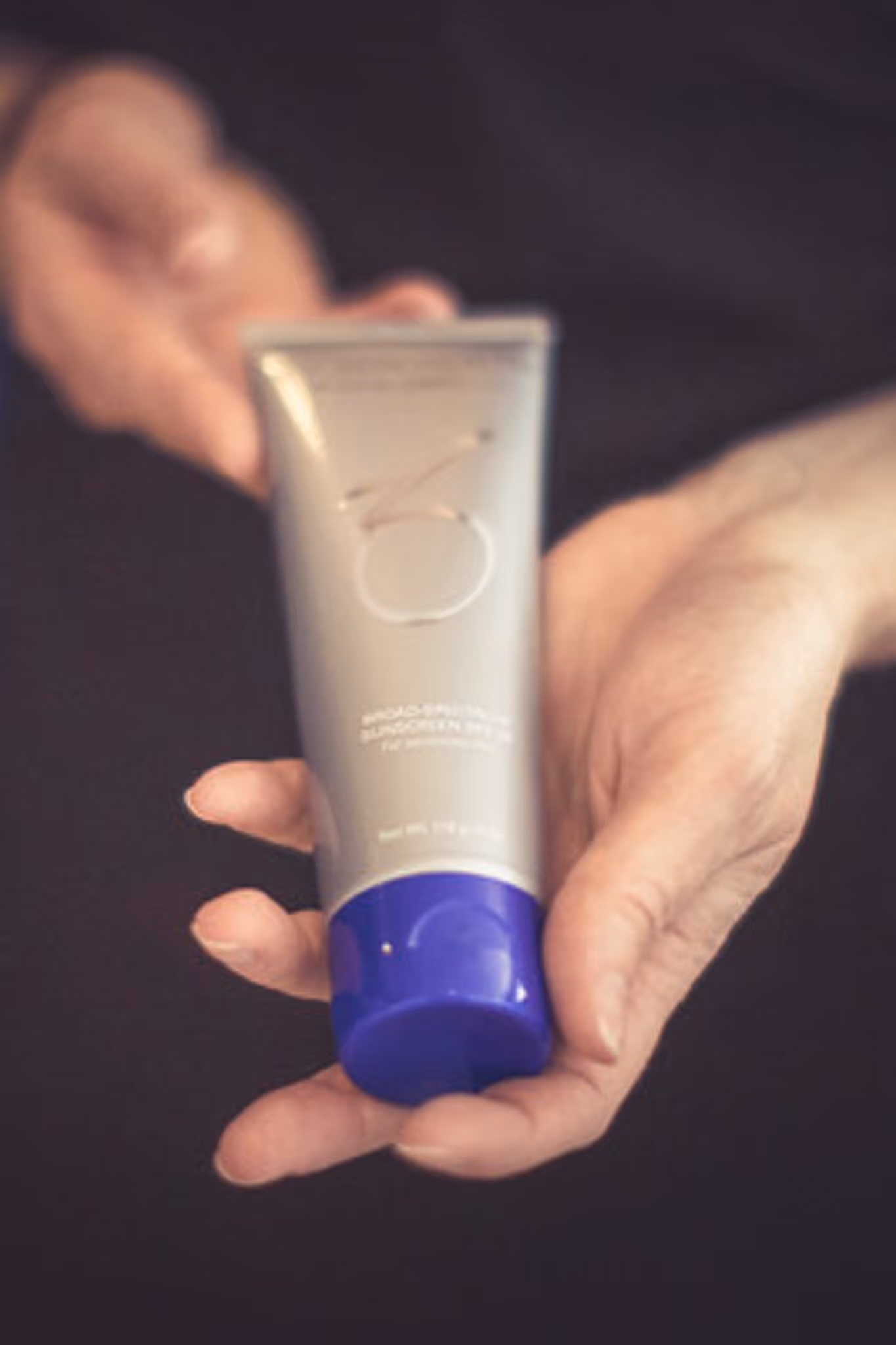 En person holder en sølvfarget tube med blått lokk i hendene, mot en mørk bakgrunn. Tuben er et hudpleieprodukt fra ZO Skin Health.