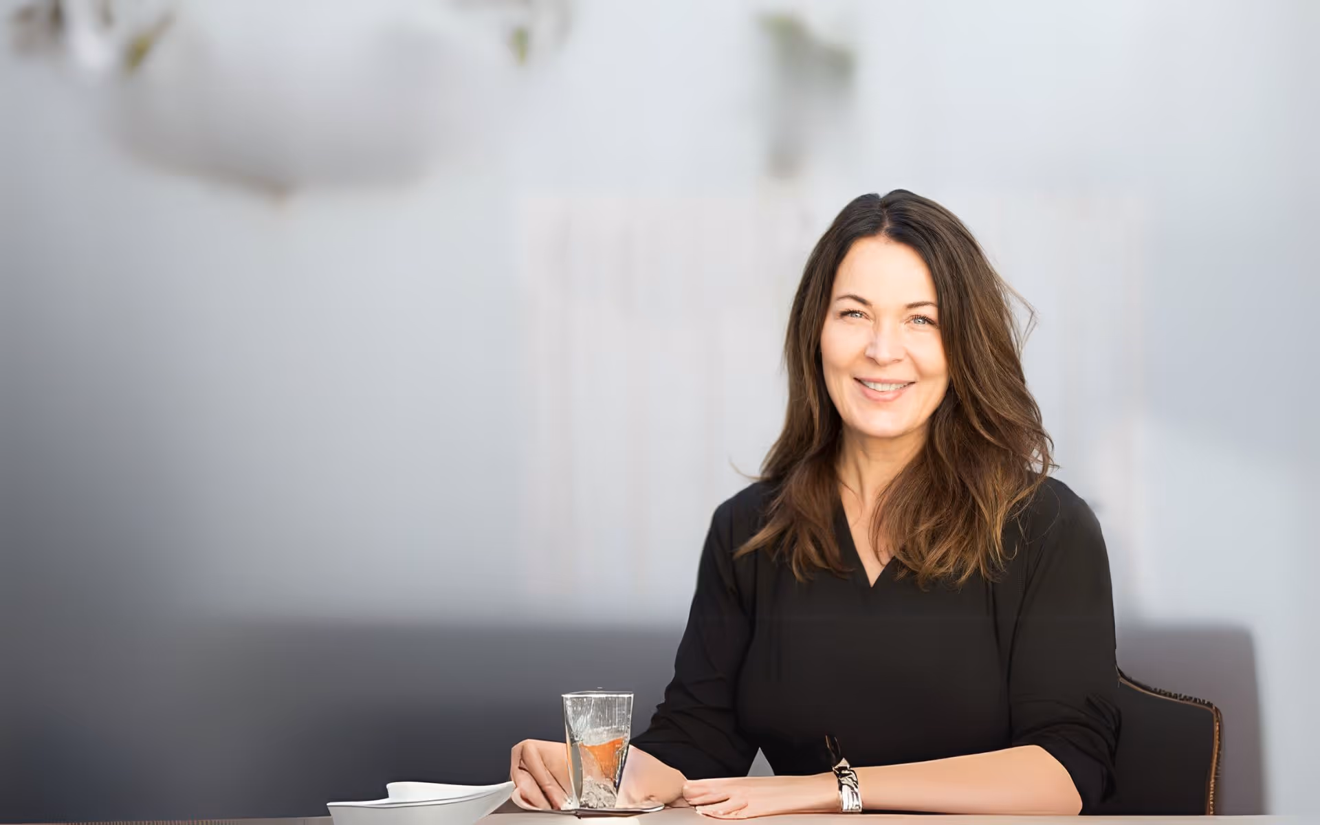 En kvinne med langt, brunt hår og svart topp sitter ved et bord med et glass vann, en skål og et fat, og smiler til kameraet i en svakt opplyst, minimalistisk setting. Kvinnen er Rannveig Welle, eier av Ad Fonte Klinikken.