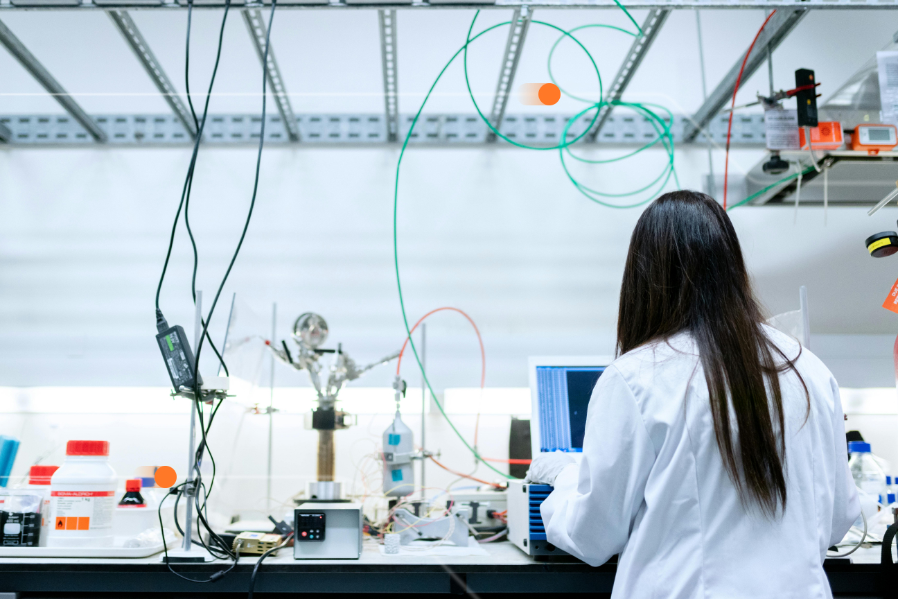 Female researcher in white lab coat working at laboratory bench with complex test equipment, including sensors, cables, and data acquisition devices in modern R&D facility