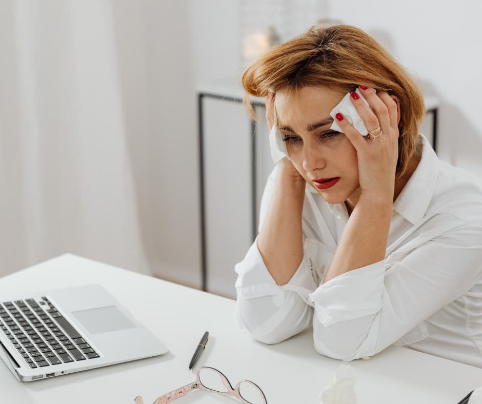 Nachdenkliche Frau mittleren Alters sitzt erschöpft am Laptop und hält sich die Hände an den Kopf – Symbolbild für Wechseljahre, emotionale Belastung, Brain Fog und Erschöpfung.
