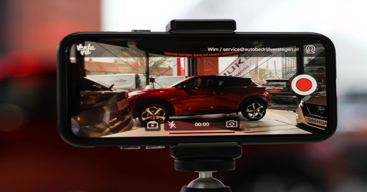Sales rep in car showroom holding phone filming a vehicle walkaround — automotive video messaging for dealerships
