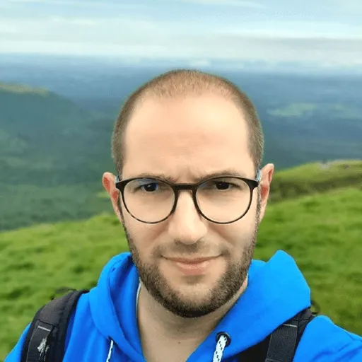 A man in a blue hoodie stands on a grassy hill with a scenic mountain view.