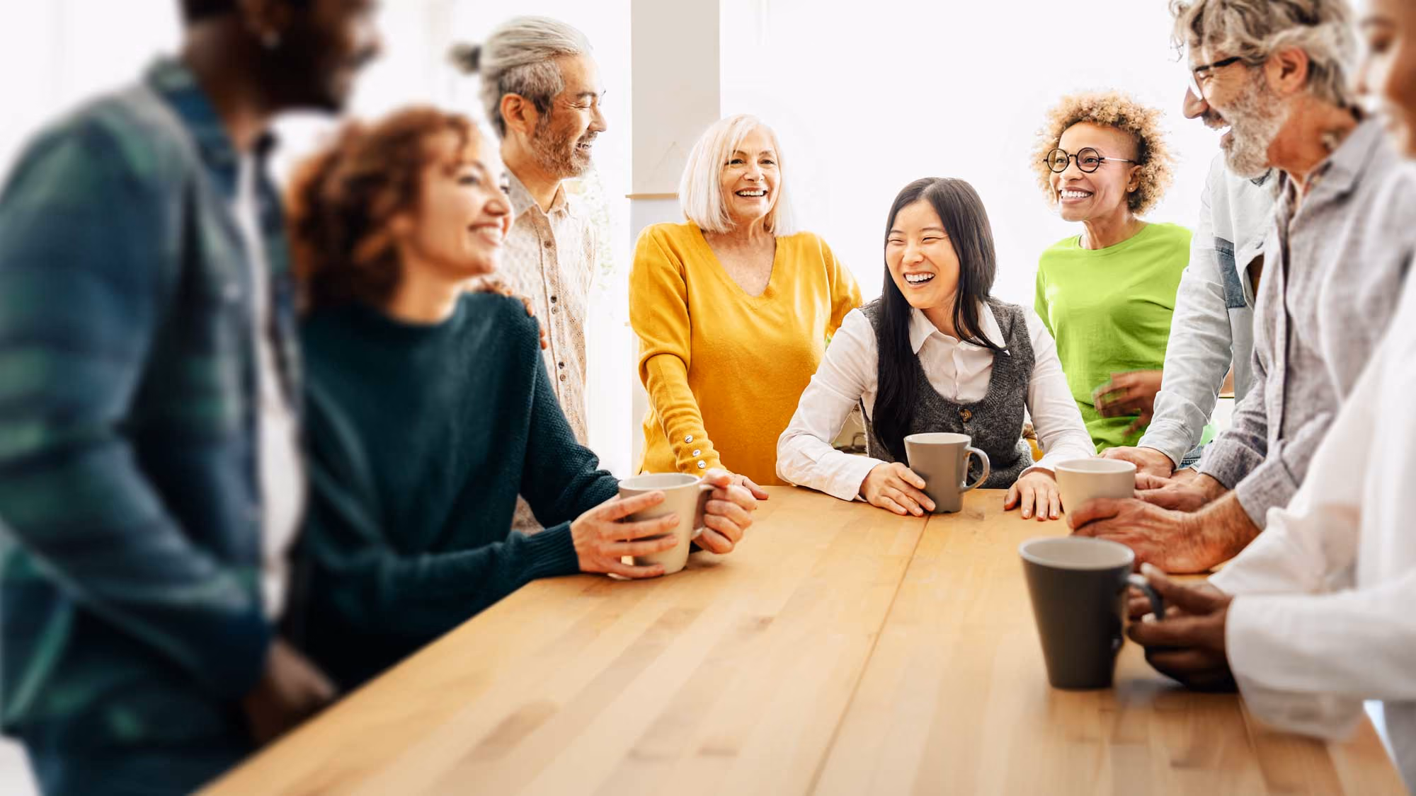 A diverse group of adults gathered around a wooden table, smiling and holding coffee mugs in a bright, casual setting.