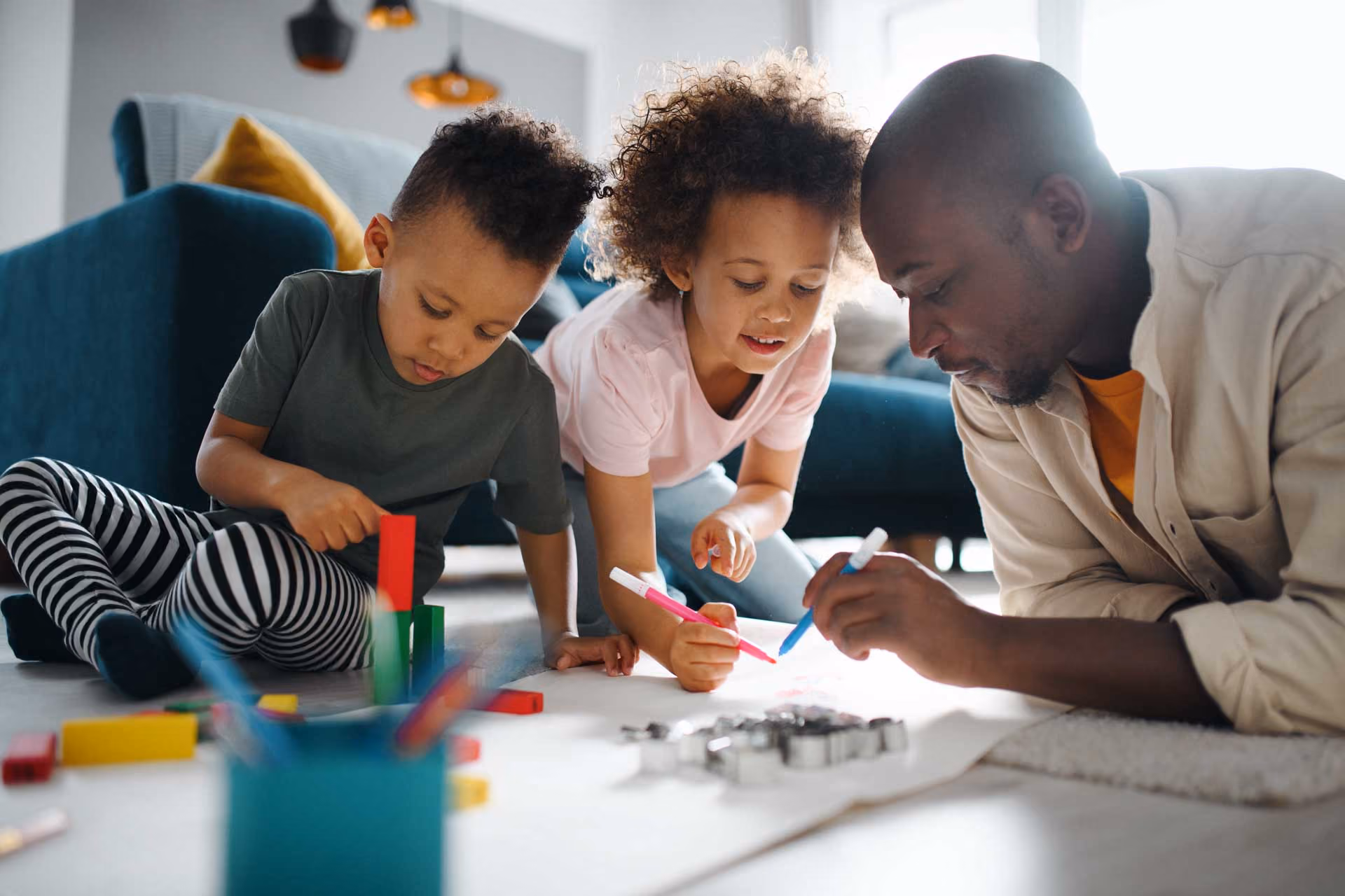 A man and two children are lying on the floor in a living room engaged in creative activities with colored markers and blocks, smiling and focused.