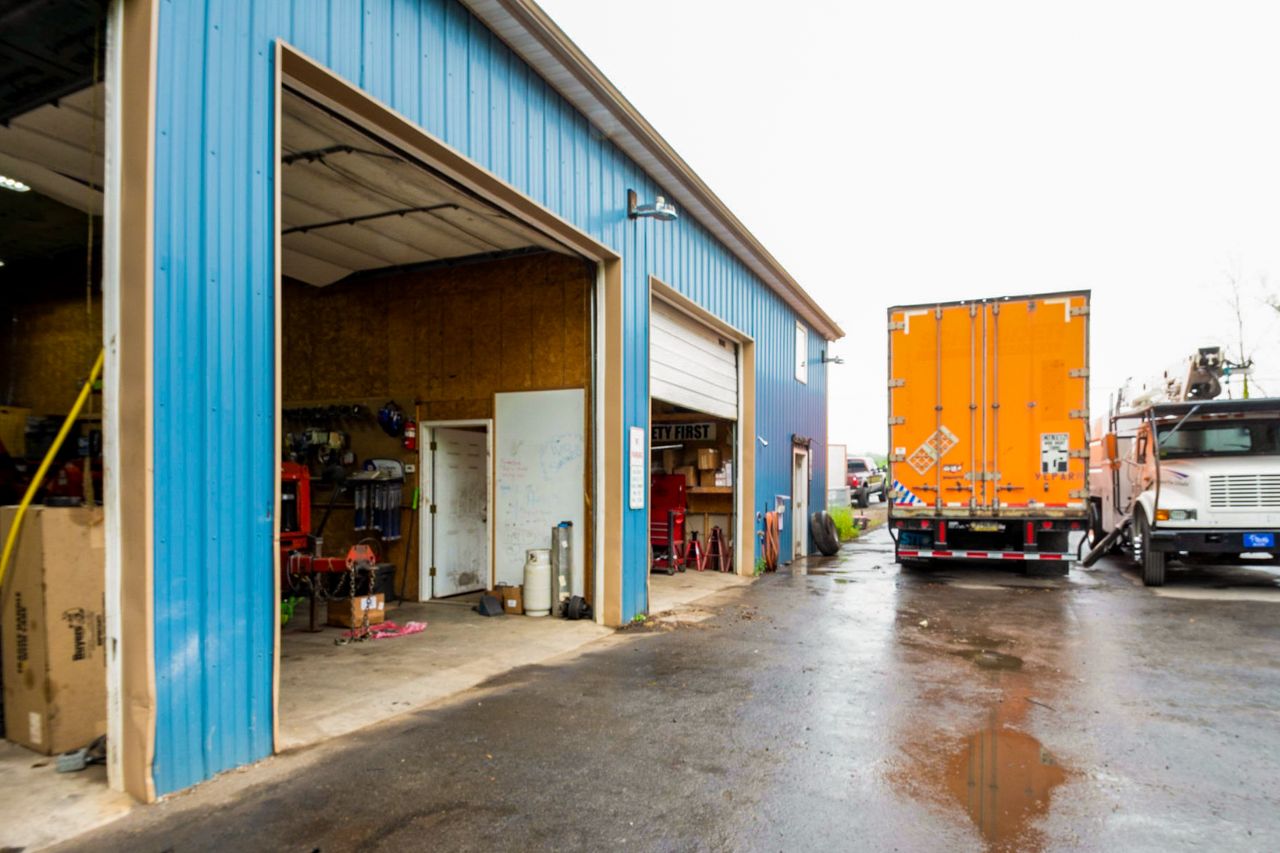 Blue metal repair shop exterior with open garage doors, visible tools inside, and an orange semi-trailer backed up outside on wet pavement.