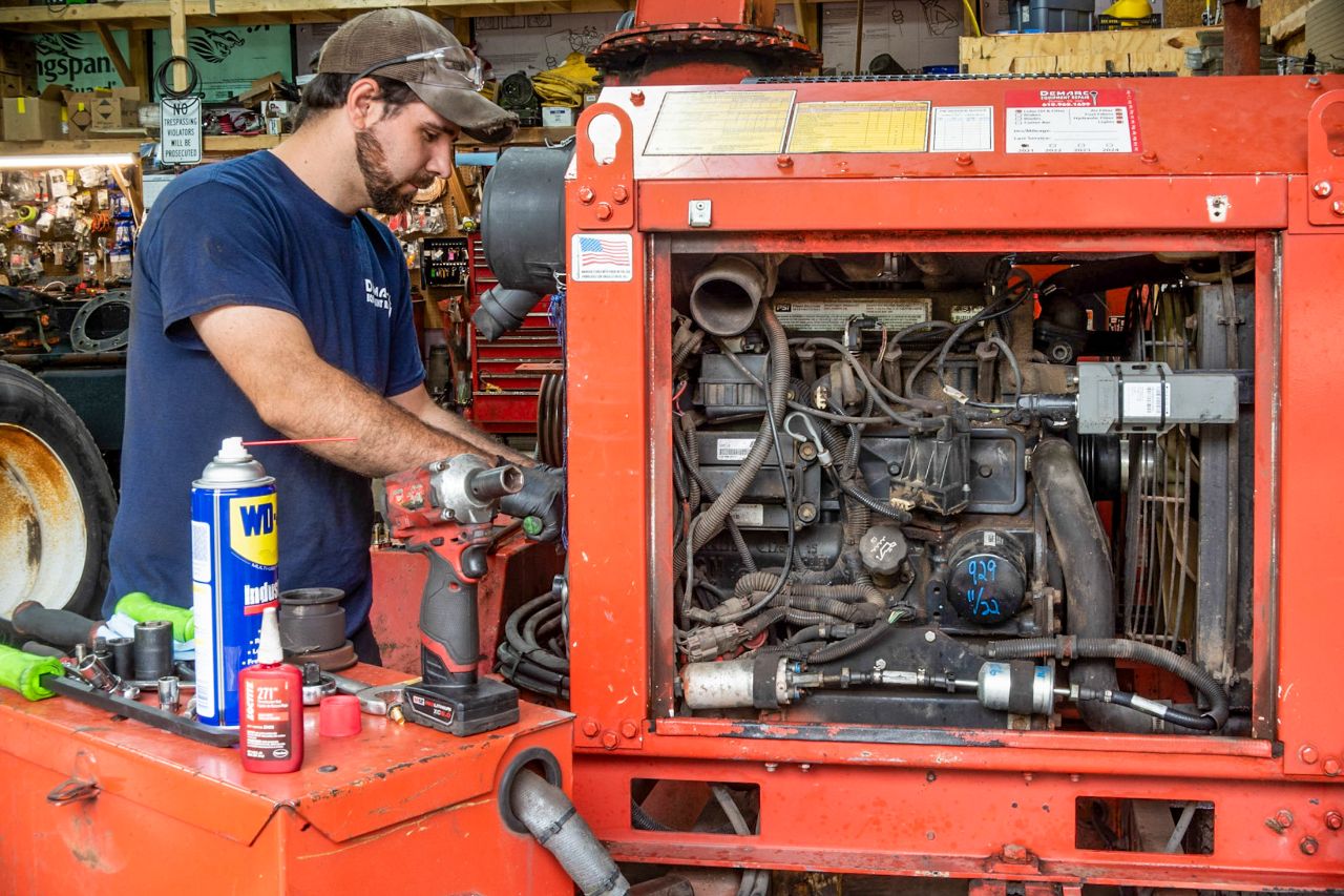 Diesel mechanic repairing engine inside red equipment housing, using power tools and lubricants, with WD-40 visible on workbench.