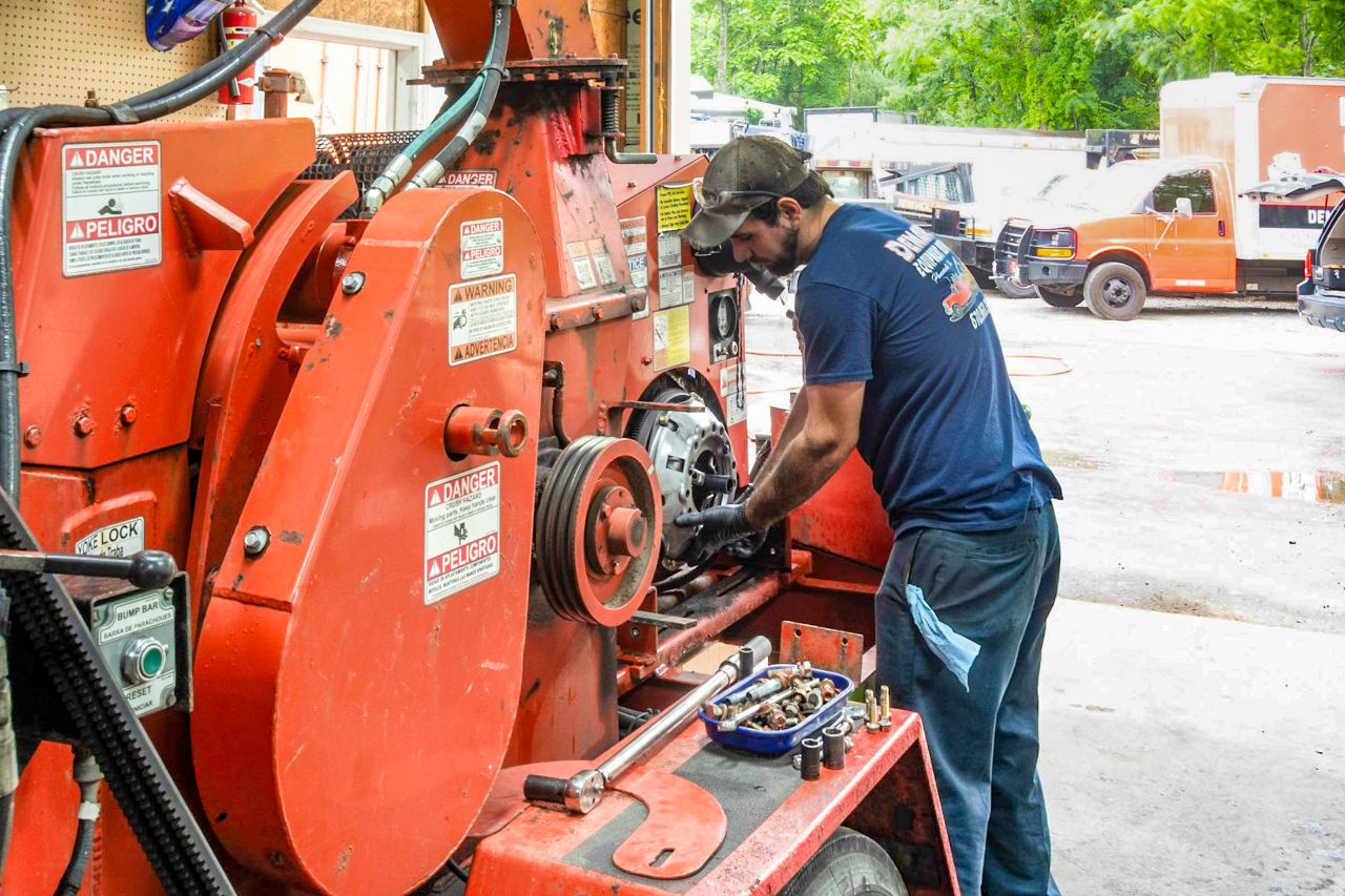 Mechanic works on large industrial orange machine outdoors, using tools and inspecting components while performing detailed equipment repair at a busy service facility