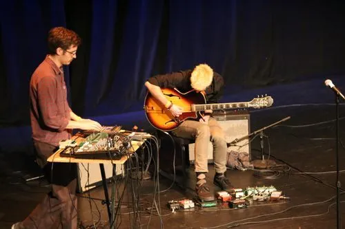 Two musicians on stage performing at an OME concert. The left one is using several electronics devices and the right one is playing electric guitar.