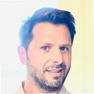 Portrait of a man with short dark hair and a beard smiling against a light background.