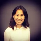 Smiling woman with shoulder-length dark hair wearing a white top against a dark blurred background.