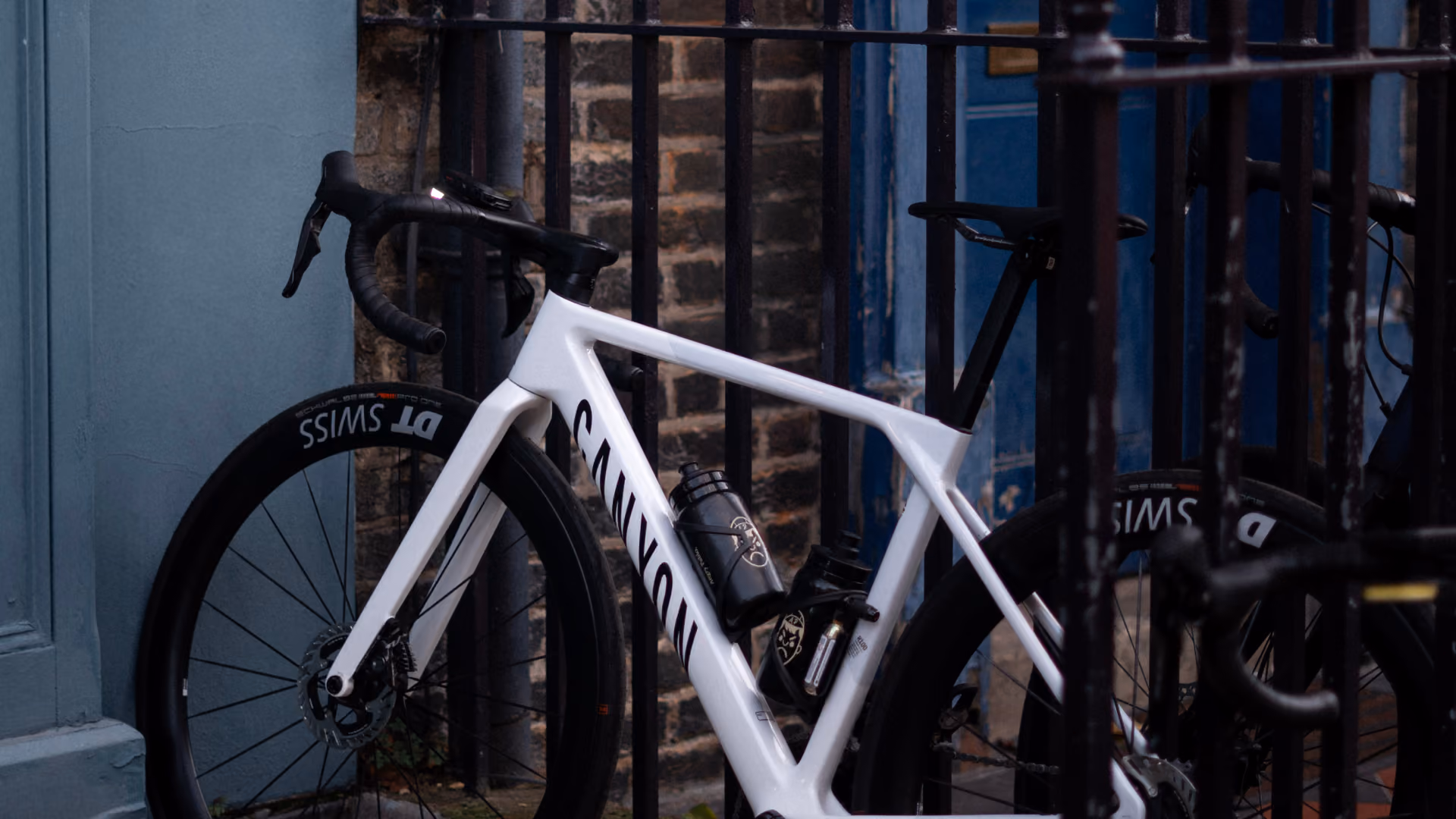 White Canyon road bike with deep-section wheels parked behind black railings outside a brick building with two water bottles attached to the frame.